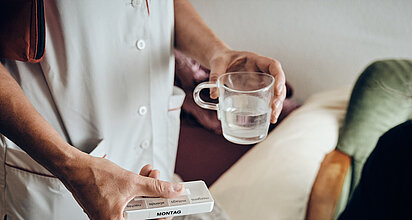 Pflegekraft mit Medikamenten in der einen Hand und einem Glas Wasser in der anderen Hand