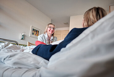 Eine Seelsorgerin sitzt bei einer Patientin am Krankenbett. Die Seelsorgerin ist gut zu erkennen, die Patientin ist nur teilweise und nur von hinten zu sehen. 