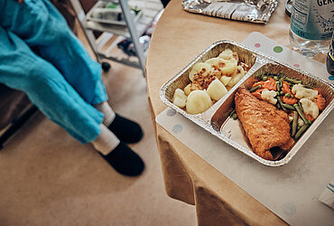 Ein Essen in einem Warmhaltebehältnis steht auf dem Tisch. Eine Frau in Hausschuhen nähert sich.  