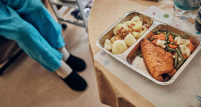 Ein Essen in einem Warmhaltebehältnis steht auf dem Tisch. Eine Frau in Hausschuhen nähert sich.  