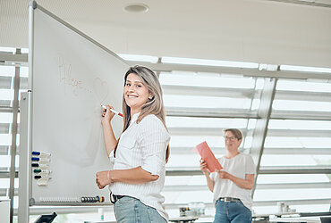 Eine Mitarbeiterin der Katholischen Sozialstation steht mit einem Stift in der Hand vor einem Whiteboard, eine Kollegin ist im Hintergrund zu sehen.
