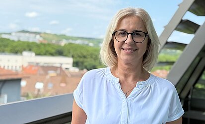 Das Bild zeigt eine 51 Jahre alte Frau in weißer Bluse auf der Dachterrasse des kirchlichen Verwaltungszentrums. Im Hintergrund sind Dächer zu sehen. 